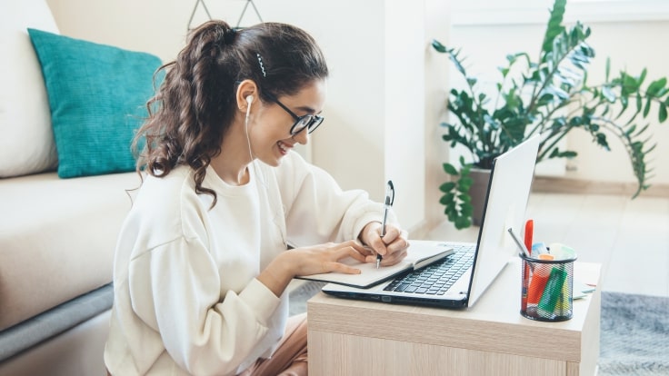 Girl in a living room, sitting on ground, in front of a computer and writing on a notebook